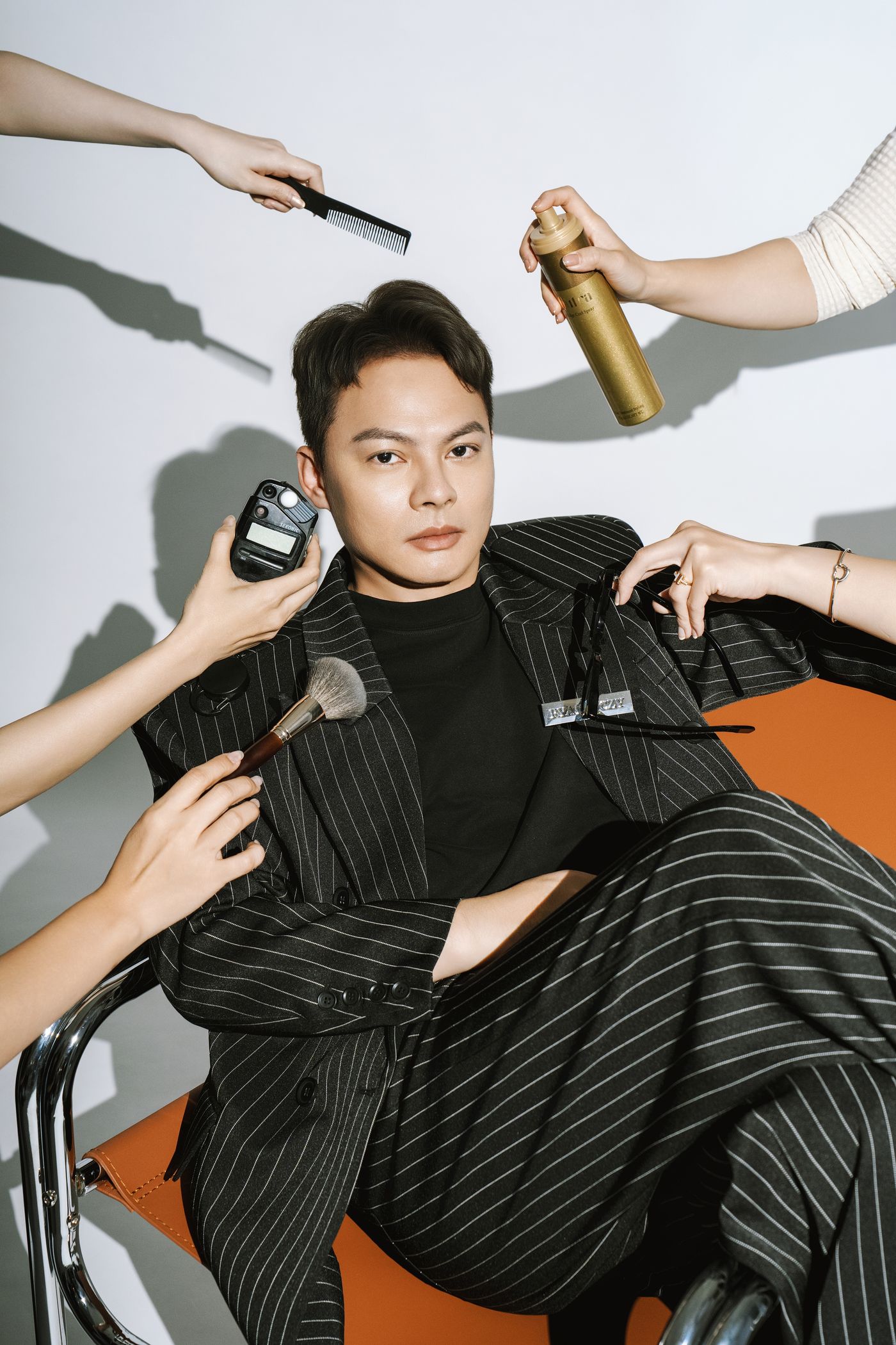白棚人物坐姿，多位造型師同時以梳子、定型噴霧、粉刷與測光錶在旁作業